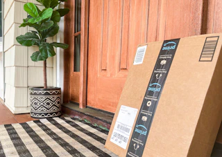 An Amazon box sitting on a front porch, leaning against the front door frame, next to a potted plant.