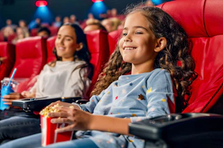 a girl holding a bag of popcorn watching a movie at the movies with her mom 