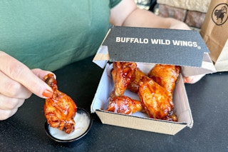 A person holding a Buffalo Wild Wing chicken wing with other chicken wings in a to go box.